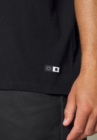 T-shirt in cotone nero con una texture liscia, caratterizzato da un piccolo etichetta con un design circolare e una stella europea vicino all'orlo. Mano parzialmente visibile.