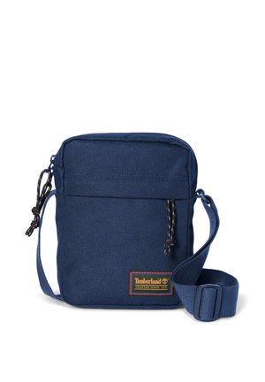 Kleine dunkelblaue Umhängetasche mit verstellbarem Riemen, vorderer Reißverschlusstasche und Timberland-Logo-Aufnäher in Orange und Grün.