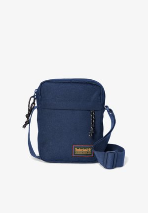 Petit sac bandoulière bleu marine avec sangle réglable, poche zippée à l'avant et écusson logo Timberland orange et vert.