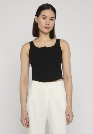 Mujer joven con cabello lacio y oscuro, lleva un top negro sin mangas con botones y pantalones blancos de talle alto, posando frente a un fondo liso.
