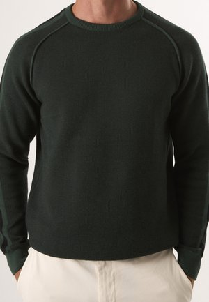 Uomo che indossa un maglione a maniche lunghe verde scuro con trama e collo rotondo e pantaloni color avorio, mani nelle tasche.