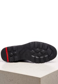 Semelle de chaussure en caoutchouc noir avec un design de bande de roulement texturé, présentant un accent rouge sur le talon. Zones renforcées pour la durabilité et l'adhérence.