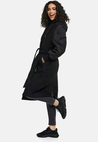 Black long coat with a hood, belted waist, and patterned fabric. Features side pockets and zippered sleeve pocket. Worn with black sneakers and gray pants.