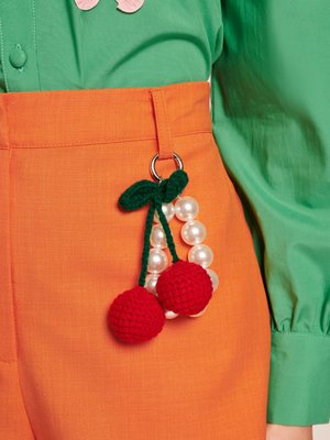 Punane lõngast kirsikujuline võtmehoidja roheliste lehtede ja valgete pärlitega aksessuaarid, kinnitatud oranžidele pükstele, seljas roheline särk.