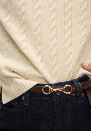 Suéter de punto de cable crema con dobladillo acanalado, combinado con jeans de mezclilla azul oscuro y un cinturón de cuero marrón con una hebilla de infinito en tono dorado.