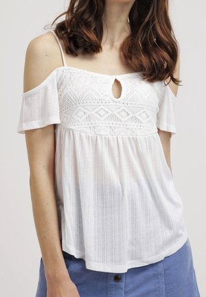 Kvinde iført en hvid cold-shoulder bluse med blonde detaljer og nøglehulssnit ved halsen, kombineret med et blåt nederdel med knapper foran.
