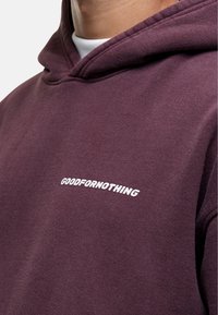 Fioletowa bluza z kapturem wykonana z miękkiego materiału, z białym logo z przodu, prezentująca luźny krój i sznurki do regulacji kaptura.