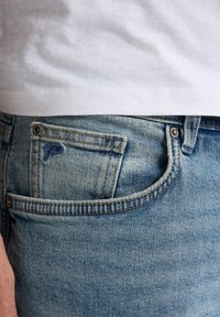 Jeans de mezclilla en azul claro con una textura desgastada, que presentan un bolsillo frontal y costuras visibles. Combinados con una camiseta blanca lisa.