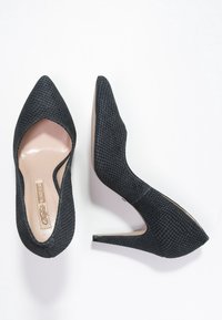 Chaussures à talons hauts noires avec une surface texturée, des pointes effilées et un design élégant. L'intérieur est d'une couleur chair lisse.