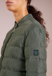 Tmavě zelená péřová bunda s vysokým límcem, horizontálním prošíváním a logem na rukávu. Má kovový zip.