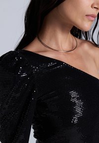 Svart paljett-top med et asymmetrisk design på den ene skulderen, med en teksturert overflate og en tettsittende silhuett. Liten metallisk halskjede som aksent.