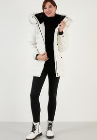 Veste blanche matelassée avec une capuche en fausse fourrure, dotée d'une doublure noire, d'une fermeture éclair et d'accents noirs, associée à des leggings noirs et des bottes blanches.