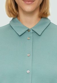 Lichtblauw polo shirt met een ronde kraag en vijf knopen, gemaakt van zachte stof met een gladde textuur. De knopen hebben een parelachtige finish.