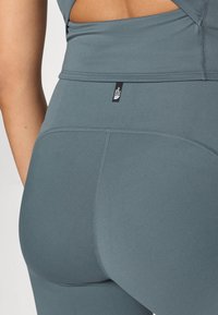 Šedé športové legíny vyrobené z elastického materiálu s hladkou textúrou, obsahujúce logo a jemné švy pre väčšiu flexibilitu.