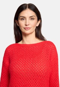 Frau mit langen dunklen Haaren, die einen leuchtend roten, strukturierten Strickpullover trägt, schaut mit neutralem Gesichtsausdruck nach vorne vor einem einfarbigen hellen Hintergrund.