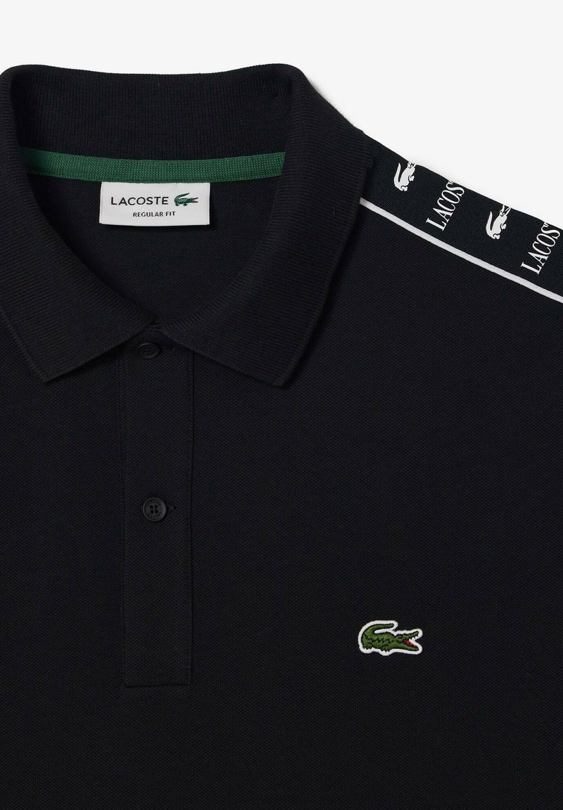 Polo negro con acento verde en el cuello. Presenta dos botones, logo bordado de cocodrilo verde y rayas laterales blancas con el nombre de la marca.