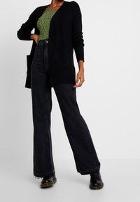 Svart stickad cardigan med öppen framsida och två fickor, ihop med höga svarta utsvängda jeans och svarta snörboots.