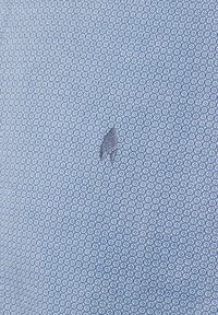 Blauer Stoff mit einem geometrischen Muster aus kleinen Sechsecken und Kreisen, ergänzt durch ein zartes, in dunklerem Blau gesticktes Logo. Weiche Textur.