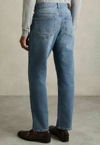 Mies, jolla on vaaleansiniset farkut ja tummanruskeat juhlakengät, seisoo neutraalia taustaa vasten selkä kameraan päin.