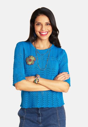 Smiling woman with long dark hair wearing a blue knit top, floral brooch, layered necklaces, colorful bracelet, and denim skirt, arms crossed.