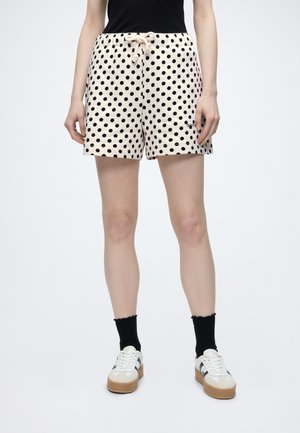 Person iført beige shorts med sorte polkaprikker, sorte sokker, hvide sneakers med beige såler og en sort top, stående på en hvid baggrund.