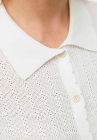 Blusa bianca a maglia con colletto scallop, chiusura a bottoni e una texture traspirante a motivo con aperture diagonali.