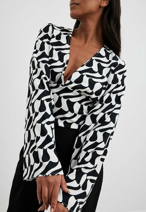 Femme portant un chemisier à manches longues à motif abstrait noir et blanc, col en V profond, posant les mains croisées devant elle.