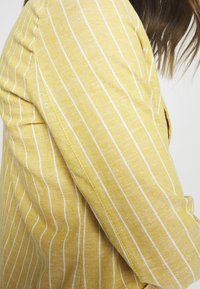 Tkanina w żółte paski z białymi liniami, z fakturowaną powierzchnią i widocznym szwem. Na zdjęciu widać bok rękawa.