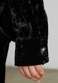 Manga de terciopelo negro con un puño que presenta una piedra preciosa decorativa acentuada en dorado. Textura suave con un brillo sutil. Vista en primer plano.