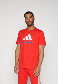 Rød bomull T-skjorte med stort hvitt Adidas-logo og "Bayern München" i blått. Korte ermer og rund hals, glatt tekstur.