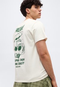 Νέος άνδρας που φοράει λευκό μπλουζάκι με πράσινο γραφικό από σάντουιτς, γυαλιά ηλίου, λεμονάδα και κείμενο «Φρέσκο! Κρατώντας τα ψωμάκια ζεστά.»