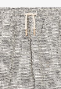 Tissu léger texturé gris clair avec un motif tissé en gris foncé et blanc. Présente une taille élastique avec un cordon de serrage et des embouts en métal.