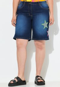 Bermudas de mezclilla en azul oscuro con costuras en contraste, que cuentan con parches de estrellas de neón en verde y rosa en la parte frontal y un dobladillo sin terminar.