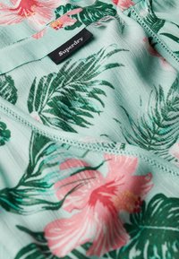 Groene bloemige stof met roze bloemen en palmbladeren, met een label met de tekst "Superdry" zichtbaar. Zachte, lichte textuur.