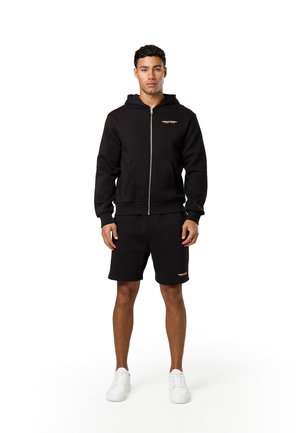 Jonge man staat naar voren gericht, draagt een zwarte hoodie met rits, zwarte korte broek en witte sneakers op een witte achtergrond.