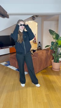 Felpa nera con zip e logo, pantaloni neri a gamba larga e sneakers bianche con plateau. Sullo sfondo si vede un divano in corduroy marrone e una pianta verde.