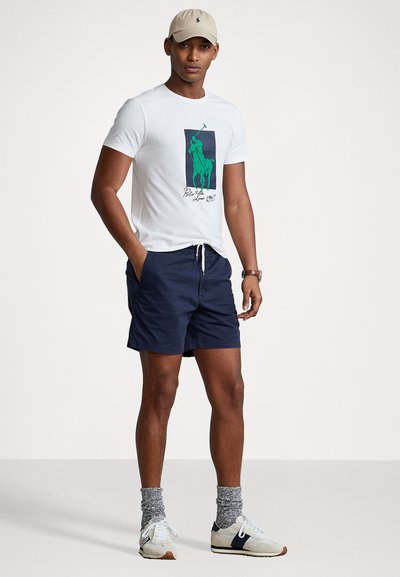 T-shirt bianco con grafica di polo verde, pantaloni corti blu scuro, cappellino beige, calze grigie, sneakers chiare con accenti scuri. Outfit semplice e casual.