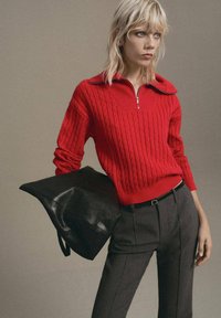 Suéter de punto rojo con cuello con cremallera, que presenta un patrón de cable texturizado. Combinado con pantalones oscuros de corte ajustado y un bolso de cuero negro.