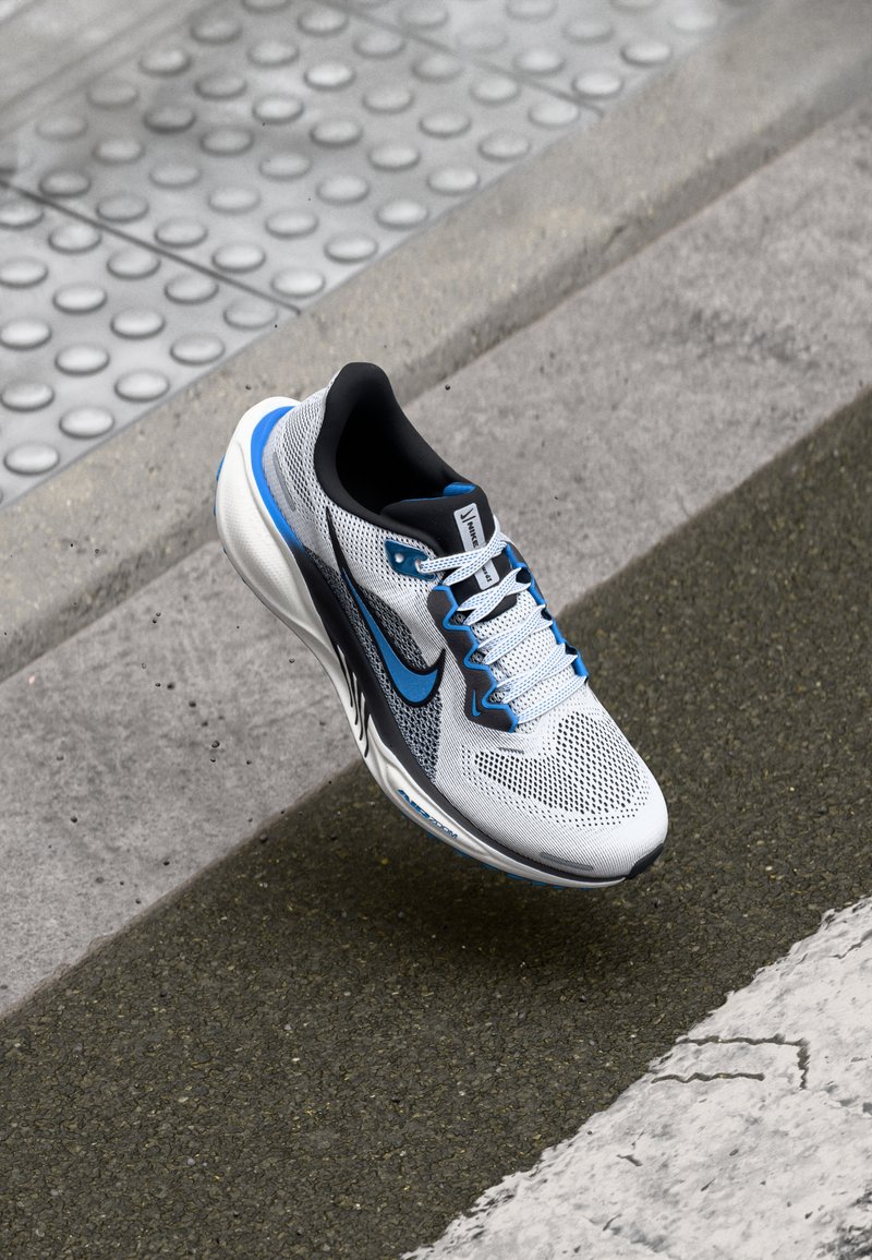 Nike tenisice za trčanje s mrežastim gornjim dijelom u bijeloj i sivoj boji, plavim akcentima, teksturiranom potplatu i vezicama. Zaobljeni vrh i dizajn s potporom.