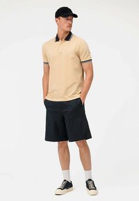 Beige poloshirt met zwart-witte accenten, gecombineerd met marineblauwe korte broek en zwarte sneakers. Model draagt een zwarte pet en witte sokken.