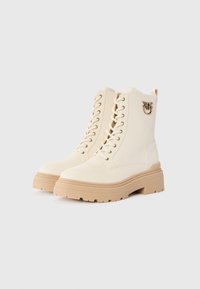 Crème-kleurige veterschoenen tot de enkel met dikke beige zolen en een kleine metalen bijversiering aan de zijkant.