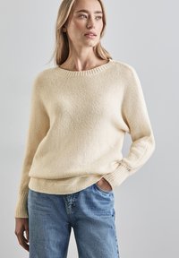 Helle beige gestrickte Pullover mit rundem Halsausschnitt, gerippten Bündchen und lockerer Passform, kombiniert mit mittelblauen Jeans.