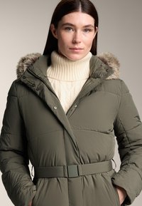 Zimný kabát olivovej farby s viazaním v páse, veľkou kapucňou podšitou umelou kožušinou a textúrovaným smotanovým rolákom pod ním.