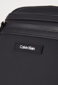 Borsa sintetica nera con superficie liscia, caratterizzata da un logo Calvin Klein in rilievo. Il design include due scomparti con zip e dettagli di cucitura discreti.