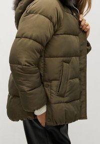 Oliwkowa pikowana kurtka puchowa z bocznymi kieszeniami i zatrzaskami, noszona na białym swetrze z kapturem wykończonym futrem.