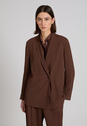 Frau mit kurzen dunklen Haaren, die einen braunen, maßgeschneiderten Blazer und passende Hose trägt, vor einem schlichten grauen Hintergrund.