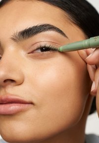 Un crayon eyeliner vert est appliqué sur la ligne des cils inférieurs. La peau du sujet semble lisse, avec un maquillage naturel mettant en valeur les yeux et les lèvres.