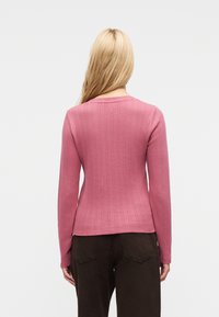 Personne blonde portant une chemise texturée rose à manches longues et un pantalon marron foncé, vue de dos sur un fond uni.
