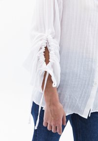 Weißes, strukturiertes Blusenoberteil mit gerafften Ärmeln und Bändern; lockere Passform. An den Füßen sind blaue Jeans aus Denim sichtbar. Lässiger Stil, leichter Stoff.