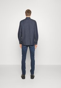 Camisa de mezclilla oscura de mangas largas, con puños abotonados y un cuello clásico, combinada con unos vaqueros ajustados azules y zapatos negros, vista desde la parte trasera.